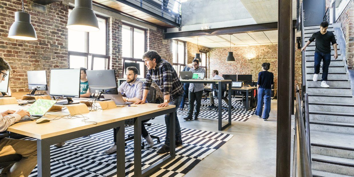 One colleague helping another at a laptop in an open-plan office